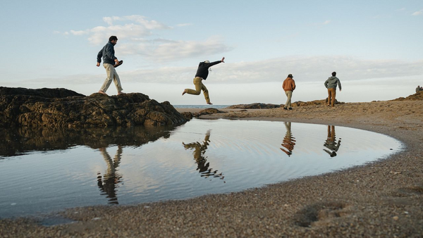 Four men in fall layers explore a rocky coastline, one jumping across a tide pool. Text reads:  "Fall Starts Here."